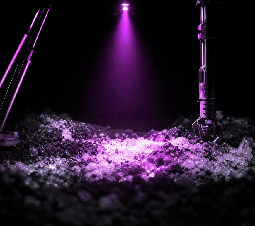 A single violet lamp beam from a lunar hopping probe lights a patch of icy regolith inside a crater otherwise in total darkness.