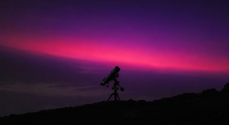 A ground telescope in silhouette on a dark hillside beneath a band of violet and magenta light spreading across the night sky.