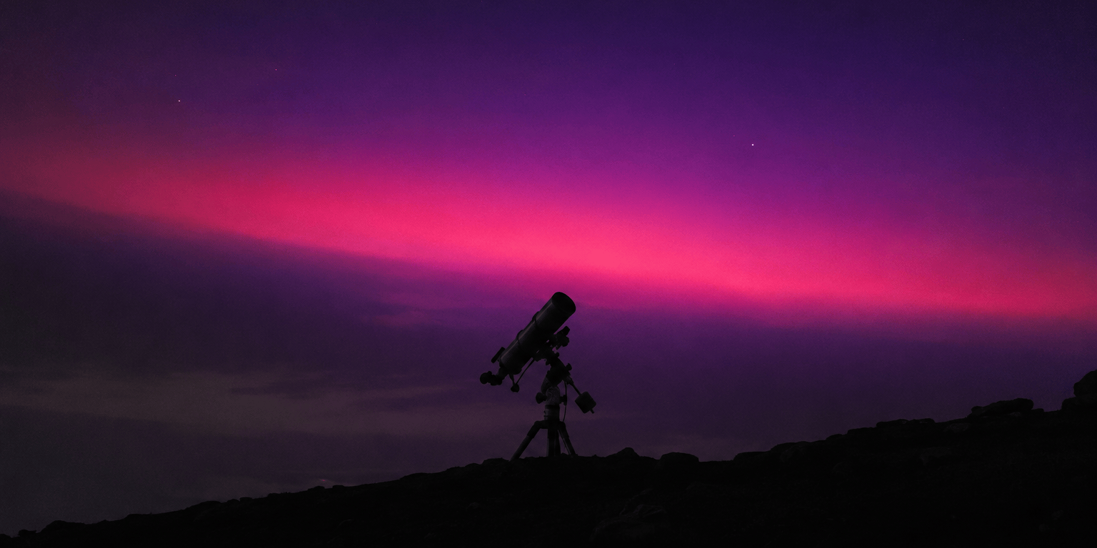 A ground telescope in silhouette on a dark hillside beneath a band of violet and magenta light spreading across the night sky.