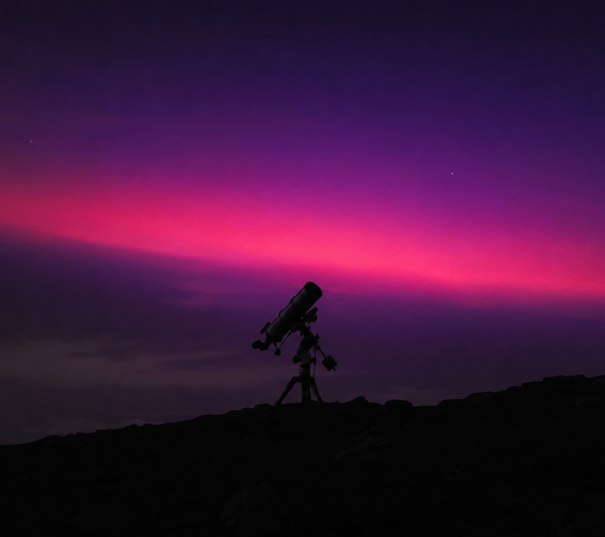 A ground telescope in silhouette on a dark hillside beneath a band of violet and magenta light spreading across the night sky.