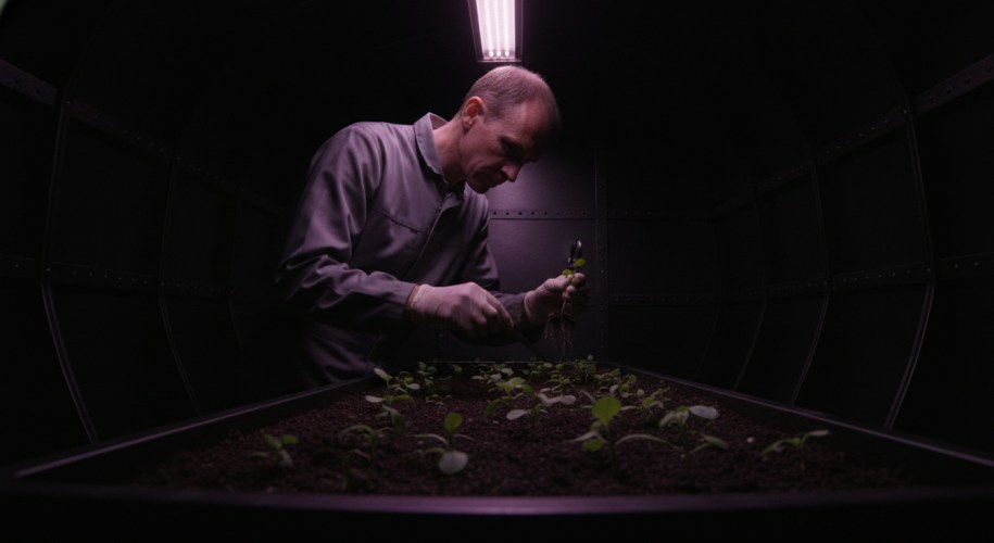 A technician in a gray coverall inspects the roots of a seedling with a hand lens above a tray of young plants in a dark greenhouse module lit by a single purple grow light.