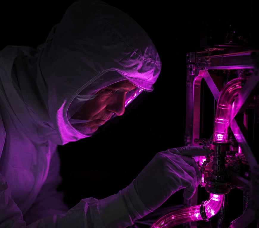 A technician in a cleanroom suit leans over a small satellite frame, a hand on a transparent fuel line glowing pink-violet in the dark.