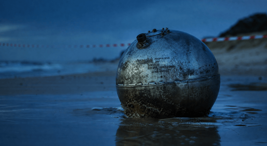 A weathered titanium propellant sphere half-buried in wet sand at the tide line of a beach at dusk, hazard tape strung along the shore behind it.