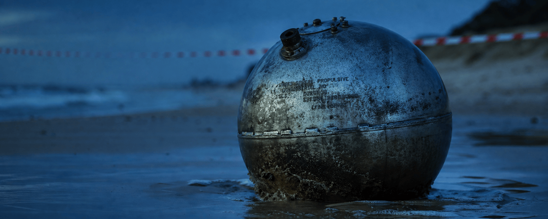 A weathered titanium propellant sphere half-buried in wet sand at the tide line of a beach at dusk, hazard tape strung along the shore behind it.