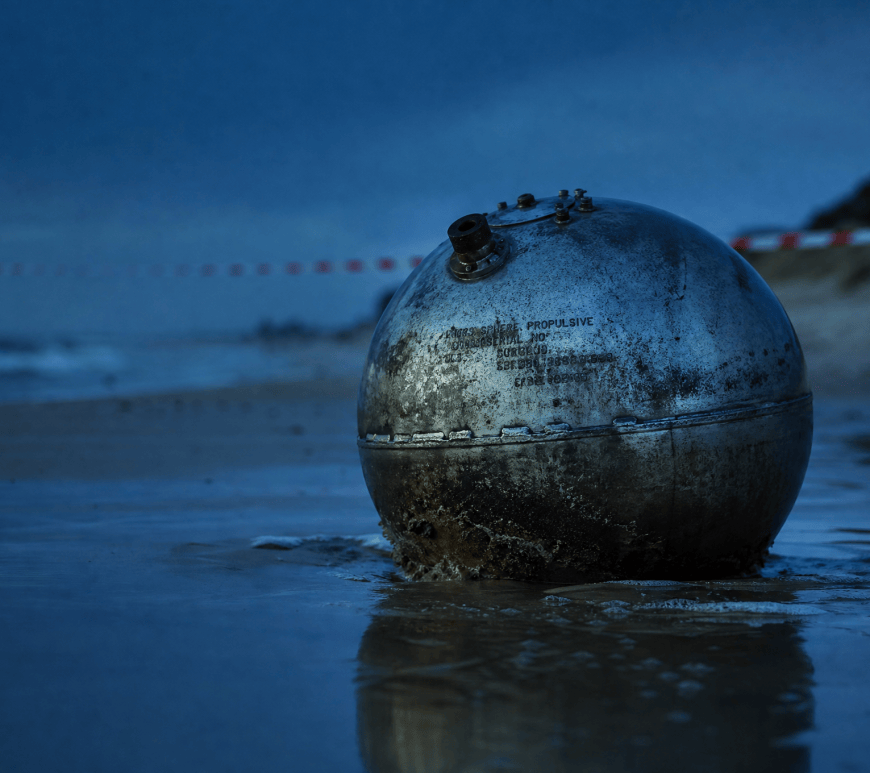 A weathered titanium propellant sphere half-buried in wet sand at the tide line of a beach at dusk, hazard tape strung along the shore behind it.