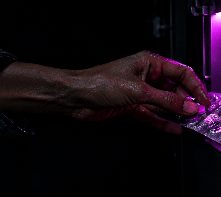 A woman's bare hand and forearm, emerging from a flight-suit cuff, press a tablet out of a silver blister pack in a dark habitat locker lit by magenta light.